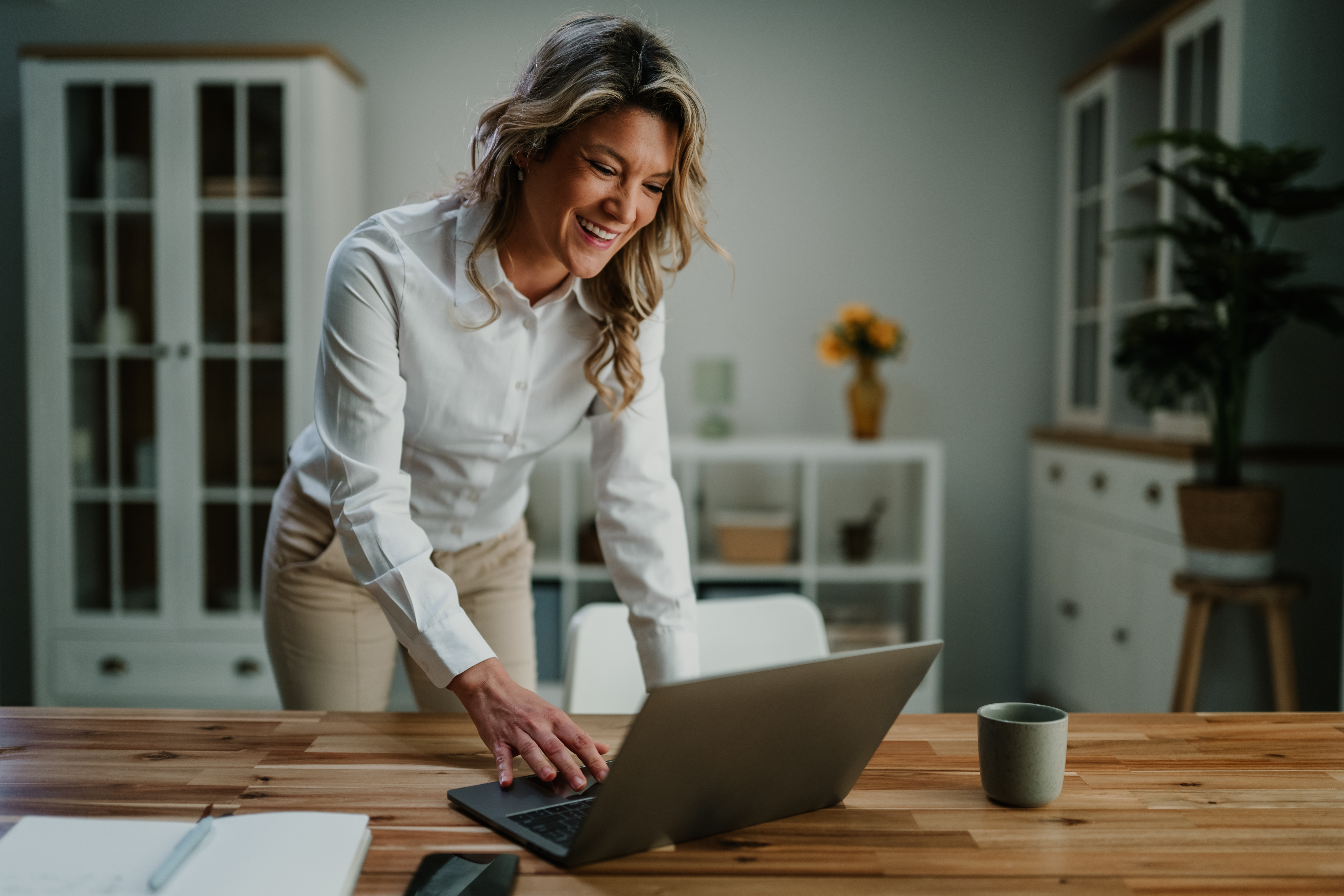 Young professional woman leaning over a laptop at a wooden desk, smiling while working from a bright, comfortable home office